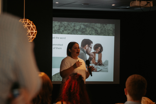 Zoe is standing at the front of a darkened room, in front of a presentation. Several people are seen listening attentively.