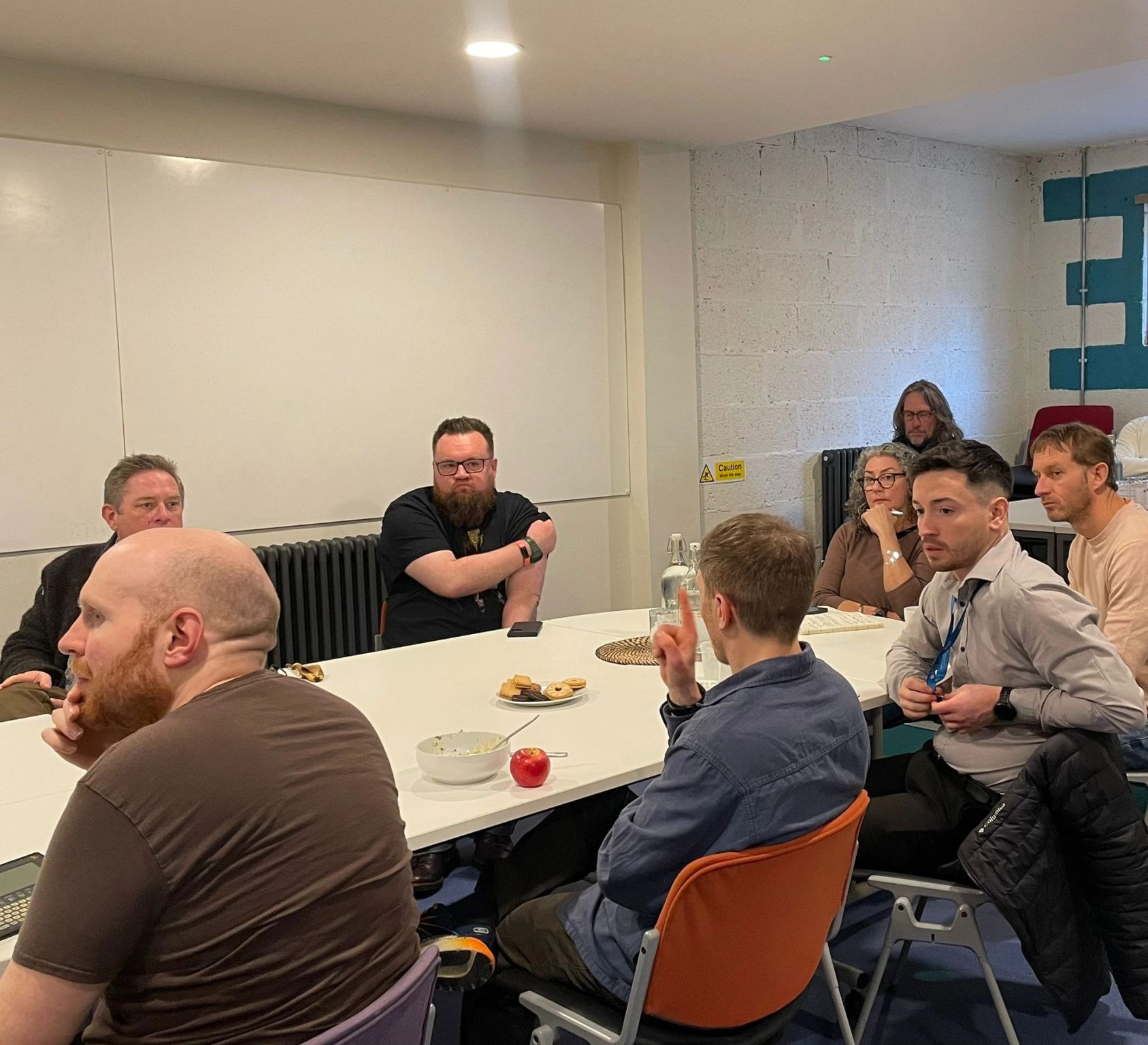 8 people sitting around a table with biscuits and water. One person is being looked at by the other