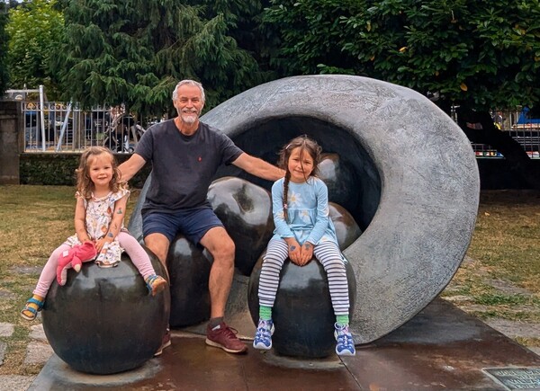 jonny and his granddaughters sitting on large bronze apples wth a massive bronze hat behind.