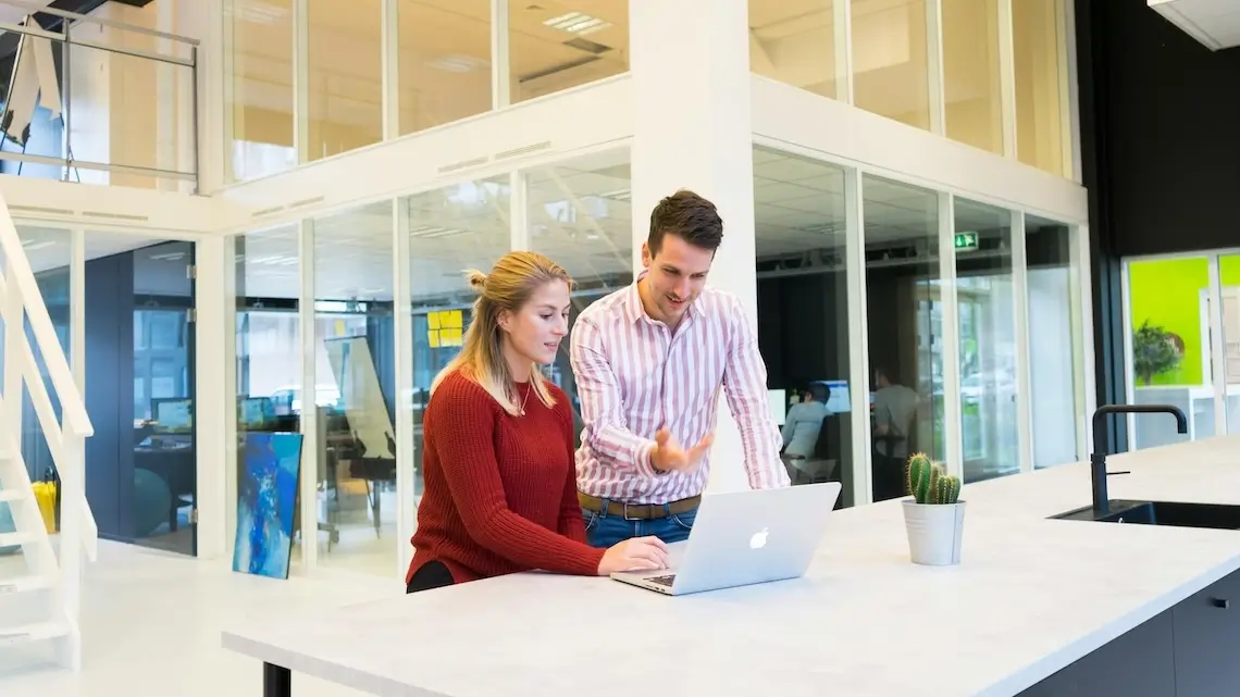 Zwei Kolleg:innen die in einem Büro an einem Tisch stehen und gemeinsam an einem Laptop arbeiten