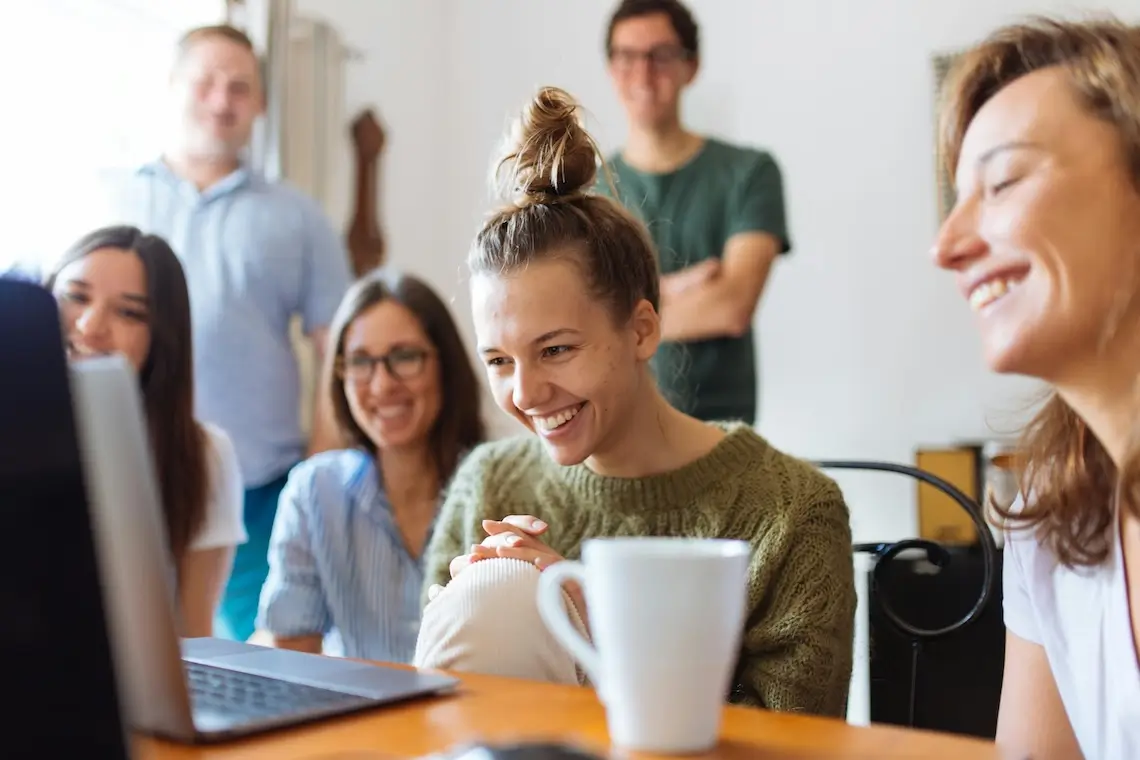 Kolleg:innen die an einem Tisch sitzen und dahinter stehen und gemeinsam auf einen Laptop schauen