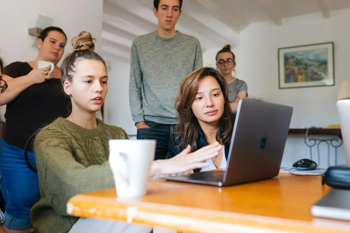 Kolleg:innen die an einem Tisch sitzen und dahinter stehen und zusammen auf einen Laptop schauen