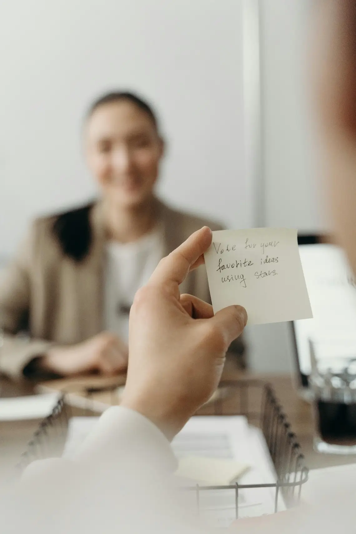 Hand hält handbeschriebenen Zettel vor der verschwommenen Silhouette eines anderen Menschen die sich gegenüber an einem Konferenztisch sitzen